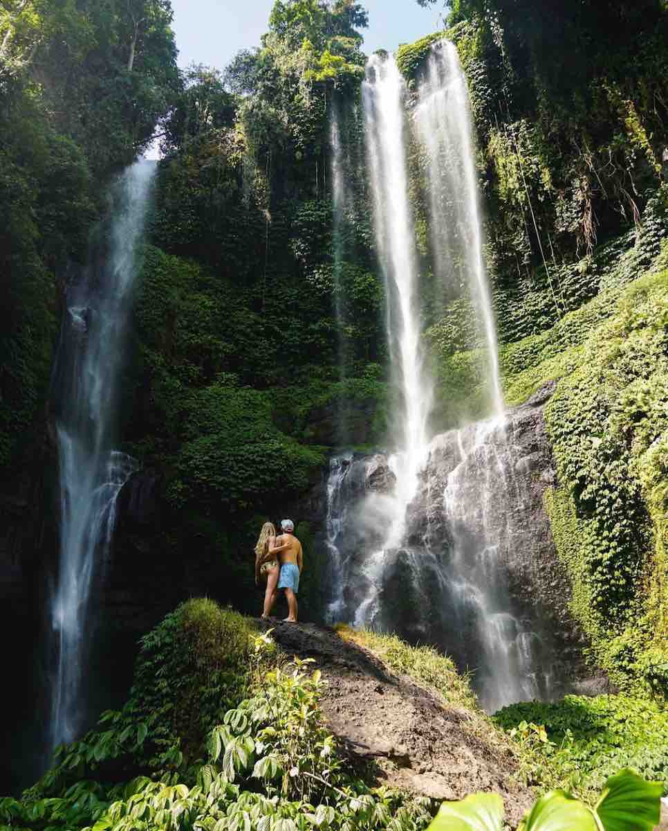 Travel couple, Cheyanne and Juan Herrera, standing in front of a waterfall in Bali, Indonesia.